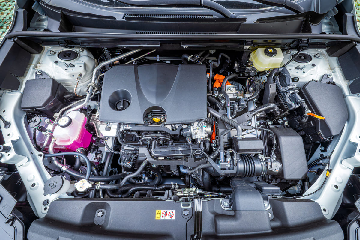 Car engine compartment with visible engine cover, hoses, coolant reservoir, and battery components.