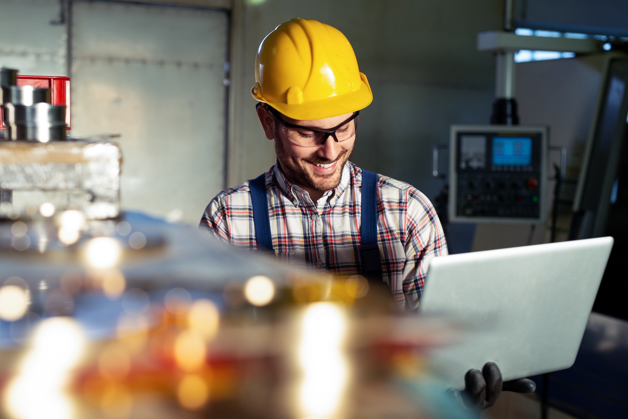 Male manufacturing technician wearing a yellow hard hat and protective eyewear smiles while viewing data on a laptop, standing near industrial machinery and metal materials.